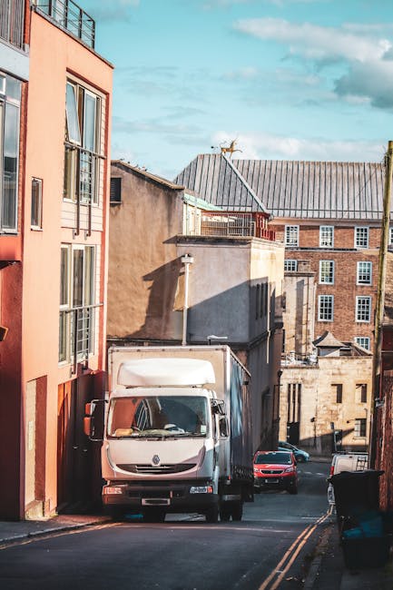 A white moving van parked on a narrow city street surrounded by multi-storey buildings with brick and stucco facades, some featuring large windows. The van is positioned adjacent to a pink residential
