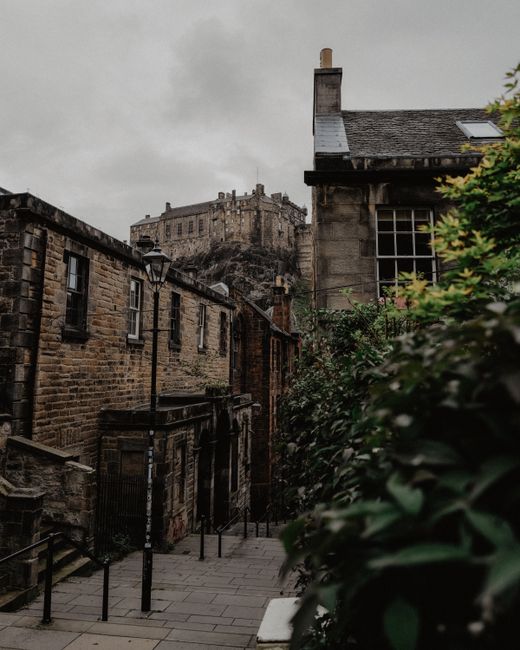 A narrow cobbled alleyway in Edinburgh's Old Town, lined with historic stone buildings featuring dark window frames and small external stairs. In the background, a large stone fortress or castle sits 