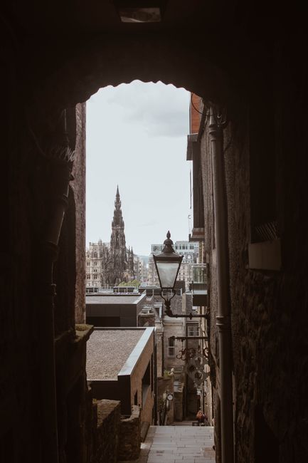 A narrow cobbled alleyway in Edinburgh's Old Town, lined with historic stone buildings featuring dark window frames and small external stairs. In the background, a large stone fortress or castle sits 
